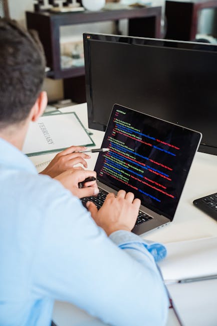 A person working at a desk outdoors, focusing on a laptop displaying lines of colourful code, surrounded by a garden setting with lush green plants, a pathway, and a hedge in the background. The works